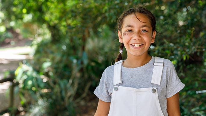 A girl is standing outside in a green, leafy environment looking happy as her parents wonder why outdoor play is important for children.