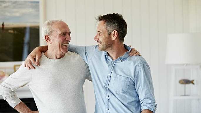 A son smiles while embracing his father as part of caring for his parents at home. 