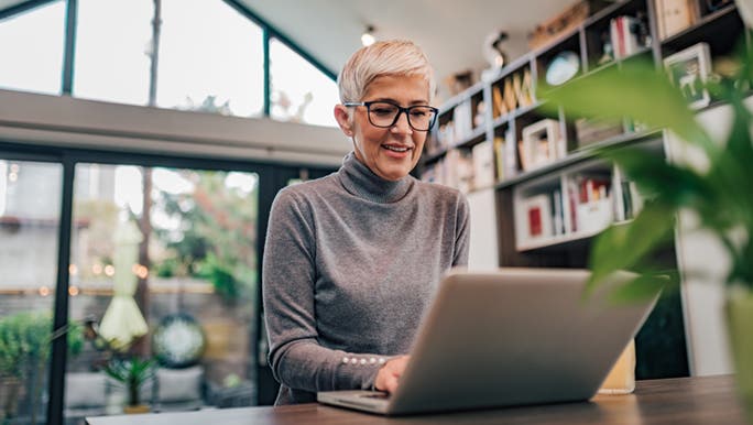 A woman is sitting at a table in a large kitchen, she is staying productive while working from home. 