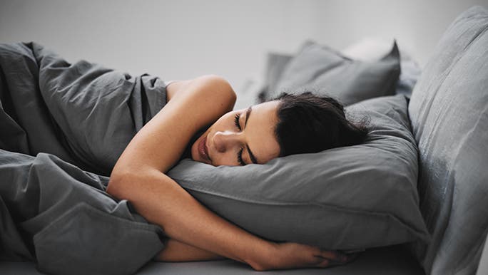 A lady hugs her pillow as she sleeps between charcoal coloured sheets. 