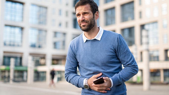 A man with dark hair and a beard wearing a blue jumper is holding his belly and looking upset, as poor gut health can impact mental health