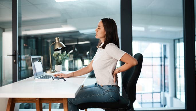 Woman sitting at her desk working and stretching out her sore lower back