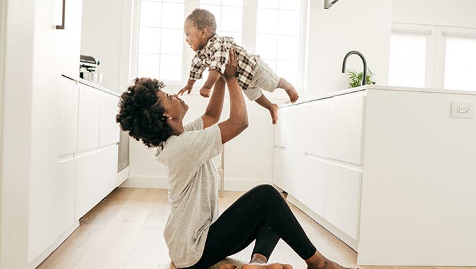 A mum is sitting on the floor of her minimalist kitchen holding her baby in the air and looks pretty happy knowing how to be a minimalist with kids. 