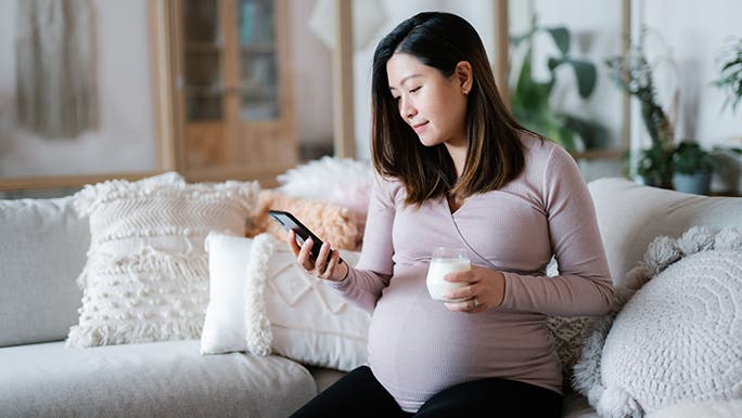 A pregnant woman sits on a couch holding a glass of milk in her hand. 