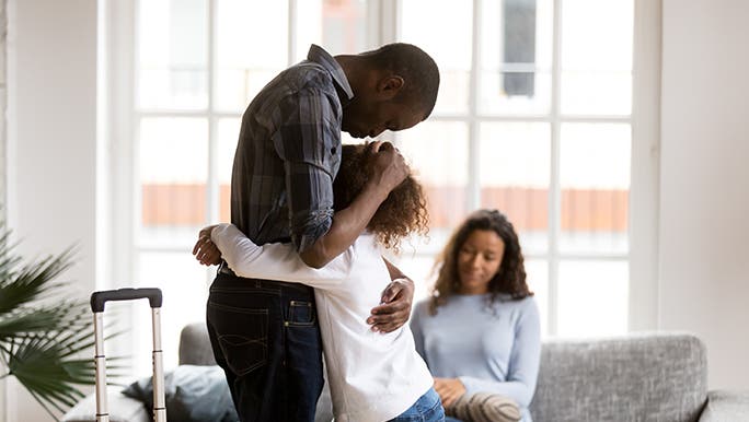 In a living room, a father comforts his daughter. The mother sits on the couch. There’s a packed suitcase waiting next to him.  