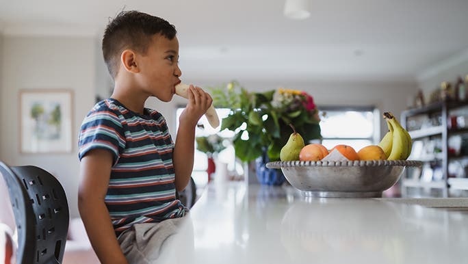 A kid is sitting at a kitchen bench eating a banana for breakfast.