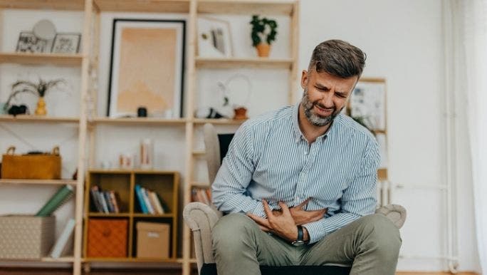 A young Caucasian man is sitting on a lounge chair and grimacing in pain as he clenches his stomach