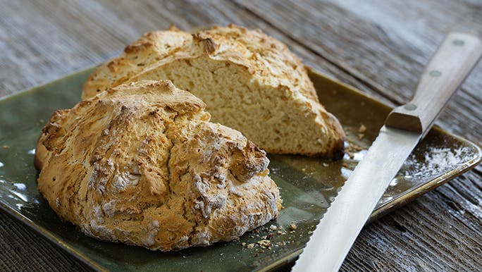 A loaf of soda bread sits on a green serving plate next to a bread knife and it could be a healthier alternative than white bread.