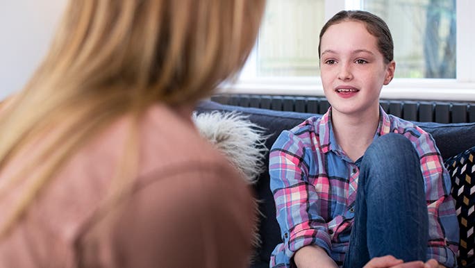 Woman talking to a tween about getting her first period at school. The girl is sitting on the couch with her arms wrapped around her knee.