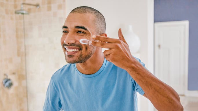 A young man wearing a blue T-shirt is smiling as he looks into a bathroom mirror and applies skin cream to his face