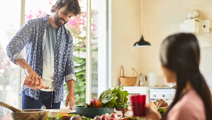 A man is standing in front of a table of fresh food and smiling as he pours salad dressing 