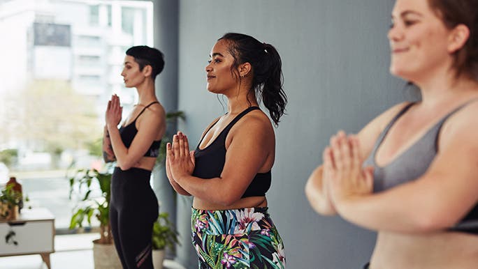Three women stand together in an exercise or yoga studio. Their hands are in prayer pose and they all look happy. 