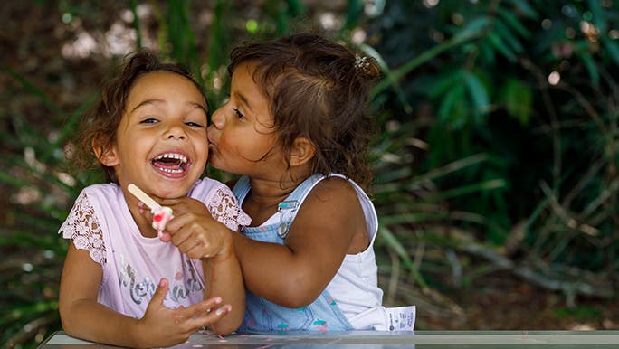 Two girls sit at a picnic table, one is kissing the other - she looks extremely grateful for the attention. 
