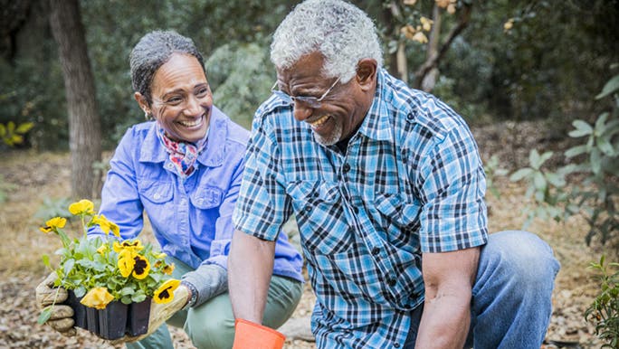 A man and woman are outside in their garden, both wearing gloves and smiling as they put  more flowers in their garden bed.