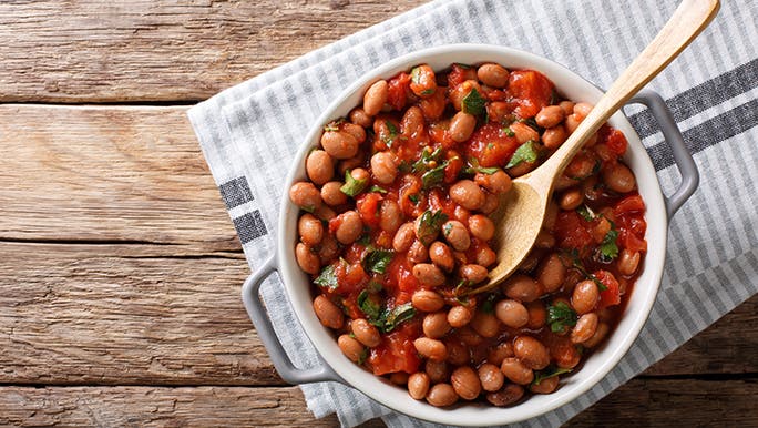 From above, a bowl of beans in tomato sauce sit on a rustic wooden table. 