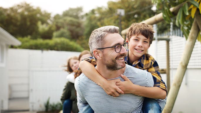 A man wearing prescription glasses piggybacks a child in a sunny backyard.