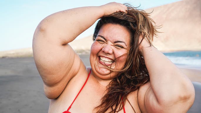 A woman in a red bikini tussles her hair on the beach, her smile is big and her eyes are closed. She knows how to feel good about herself. 