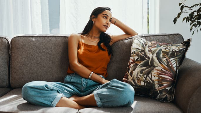 A young dark-haired woman wearing jeans and an orange top is sitting on a sofa looking fatigued