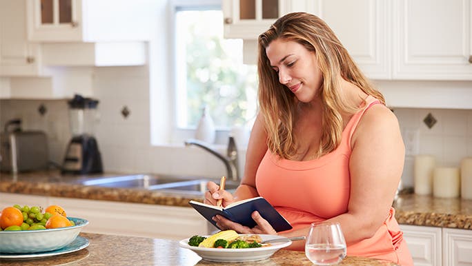 Happy woman in a coral coloured casual dress, sitting at a bench in a modern, bright kitchen. There’s a bowl of vegetables in front of her and she is taking notes in a diary about brain-healthy foods. 