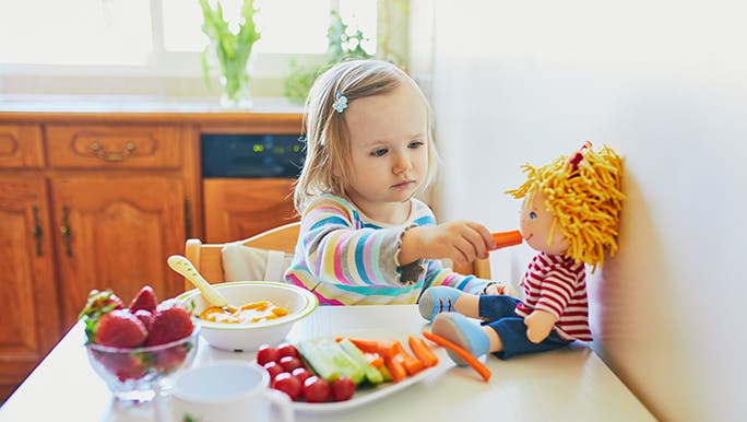 A toddler feeds a rag doll a carrot. She is sitting at a table that is packed with veggies.