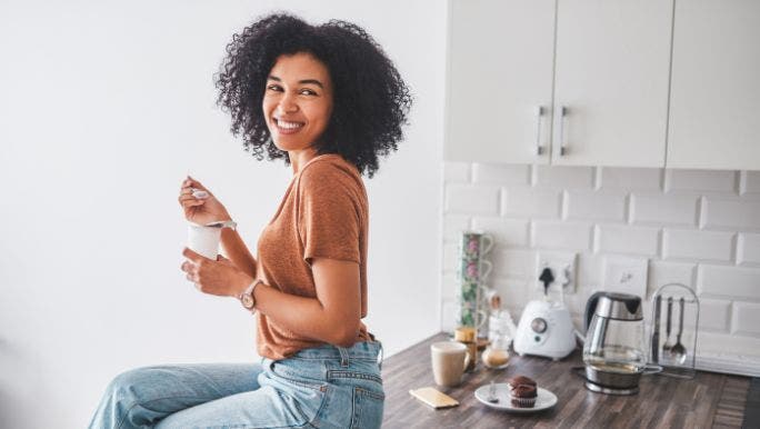 A young woman is sitting on a kitchen bench eating yoghurt