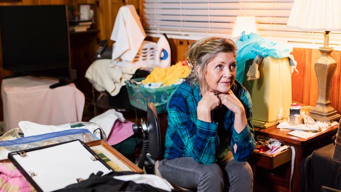 A middle-aged woman is sitting in a cluttered room cupping her chin with her hands