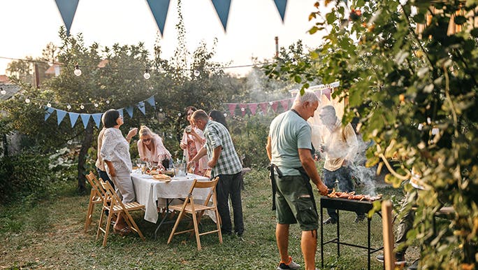 A family gathers in a leafy backyard for a BBQ. There are blue flag garlands strung around and the cook wears an apron. 