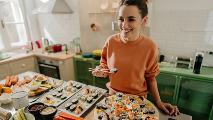 A young woman is standing in her kitchen and smiling as she eats sushi made with salmon and tuna