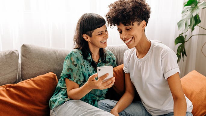 A young dark-haired woman wearing a green shirt sits on a couch holding her phone and chatting to her female partner, who has dark curly hair and is wearing a white T-shirt