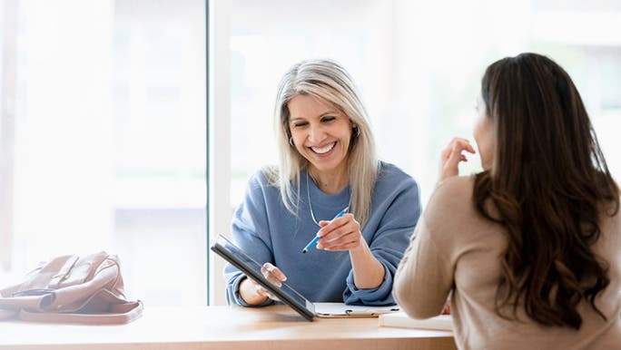 A blonde woman wearing a blue jumper is smiling and talking to a brunette woman during a medical consultation