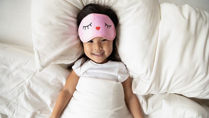 A child about to take a nap smiles while wearing a pink sleeping mask to help her get the best nap length possible. 