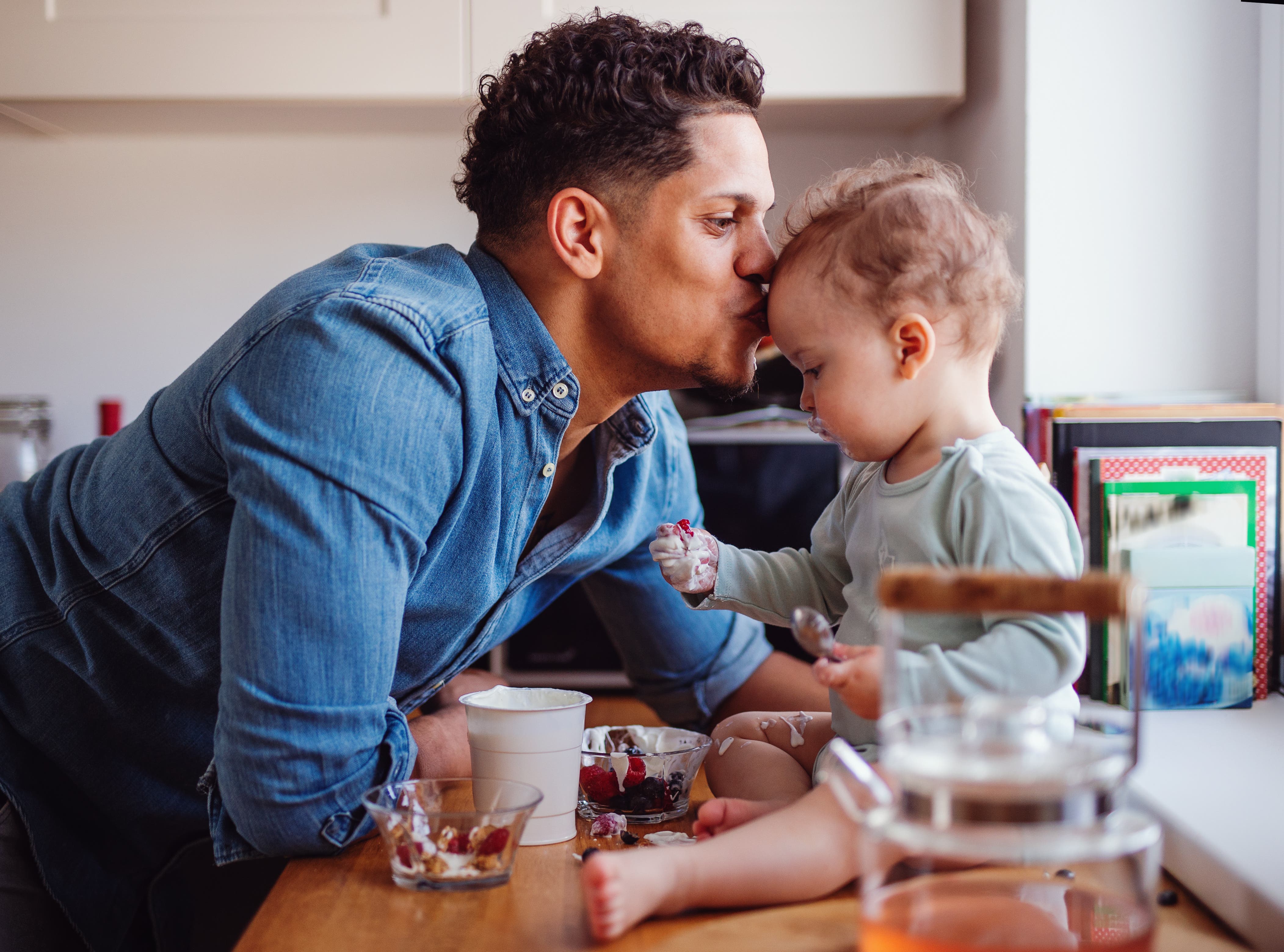 A man and a toddler are eating yoghurt and fruit in a kitchen
