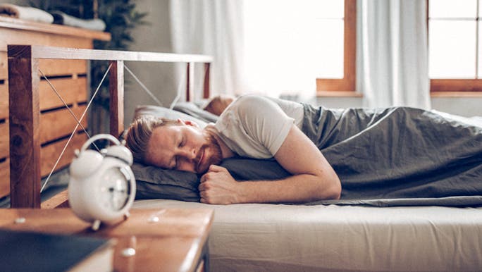 A Caucasian man with a beard and wearing a white T-shirt is sleeping in a bright room beside a white alarm clock on a bedside table