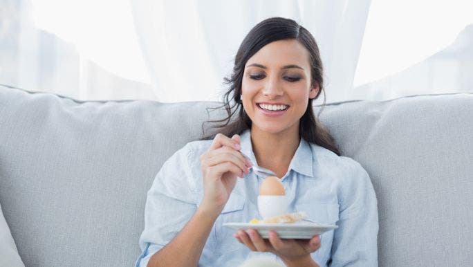 A young woman is sitting on a sofa and smiling as she eats a boiled egg