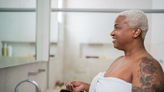 A woman wearing a white towel and with a tattooed arm is smiling as she looks at her face in a bathroom mirror before applying face cream