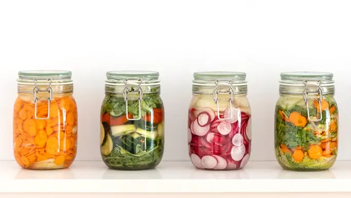 A line of picking jars full of colourful fermented vegetables is displayed in front of a white wall