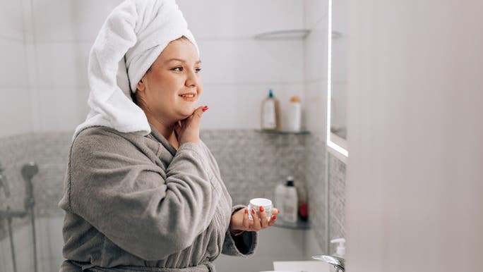 A woman wearing a bathrobe with a towel wrapped around her head is applying face cream while looking at a bathroom mirror