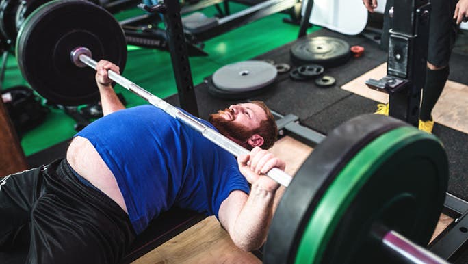 A white Caucasian man with a large belly is in a gym doing bench press exercises with heavy weights