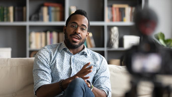 A life coach sits on the couch, he is mid sentence talking to a camera. He is wearing glasses and looks energetic and uplifting. 