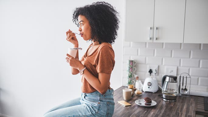 A young woman sits on her grey kitchen bench eating probiotic yoghurt at breakfast which is a good time to take probiotics.
