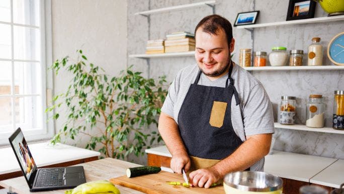 A young man is smiling and preparing fresh vegetables in a kitchen while watching a video on a laptop