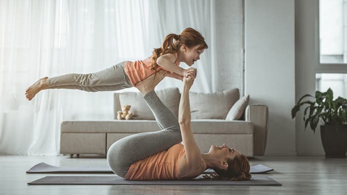 Mum is laying on the floor of her lounge room on a yoga mat. She is lifting her child in the air with her feet. 