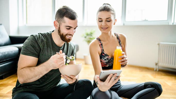 A young man and woman look at a handheld device while sitting cross-legged on the floor, while the man eats a salad and the woman drinks a juice