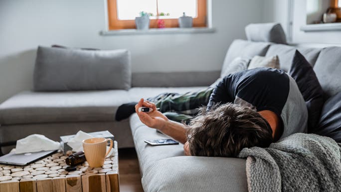 A man is lying on a grey couch, sick. Being ill can be one the reasons for low sex drive.