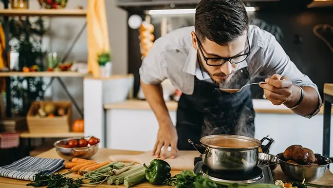 A men is bending over to taste some soup on his spoon. He is cooking in the kitchen.