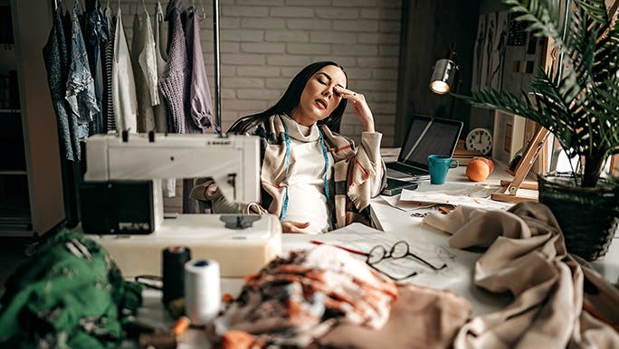 A pregnant woman sits in her workplace looking exhausted. There is a sewing machine in front of her and fabric everywhere. 