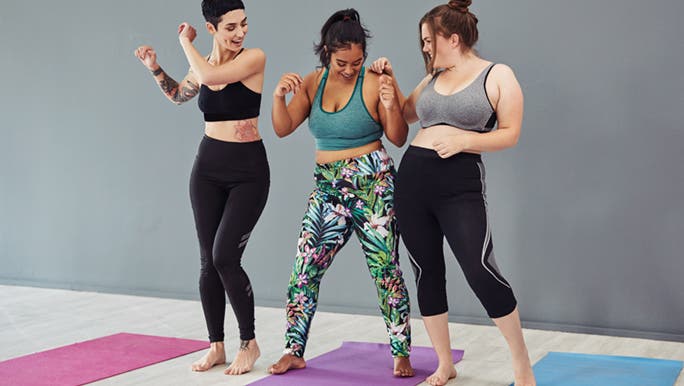 Three beautiful women of different sizes are wearing activewear on colourful yoga mats. They are dancing as a way to get motivated to lose weight.
