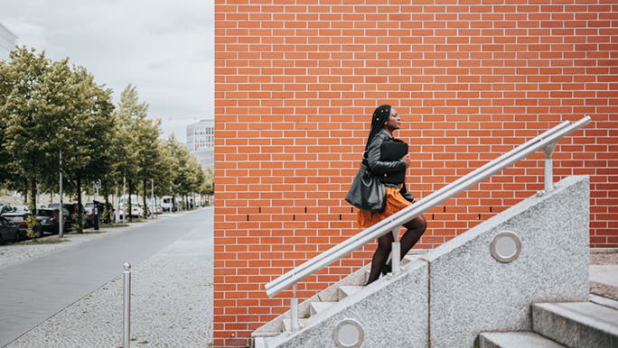 A woman is choosing to take the stairs so she can get her daily exercise in. There is an orange brick wall behind her and she looks to be in office wear. 