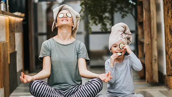 Mother and daughter are sitting down on the ground with their legs crossed. They are both wearing pjs and doing some self care with cucumbers over their eyes and a towel wrapped around their hair. 