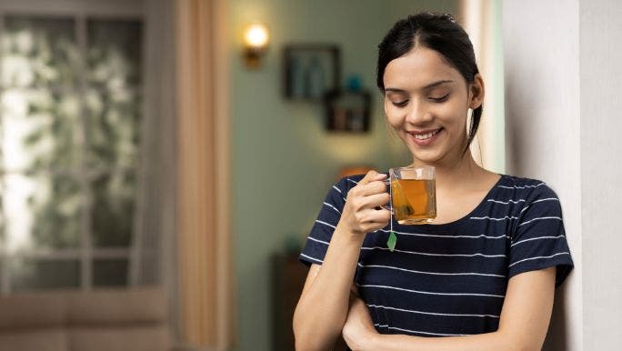 A young woman is leaning against a wall as she smiles and drinks herbal tea